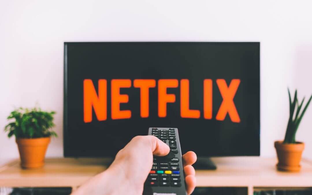 Osoba drži daljinski upravljač usmjeren prema televizoru s logotipom Netflix, okružena biljkama u loncima.
