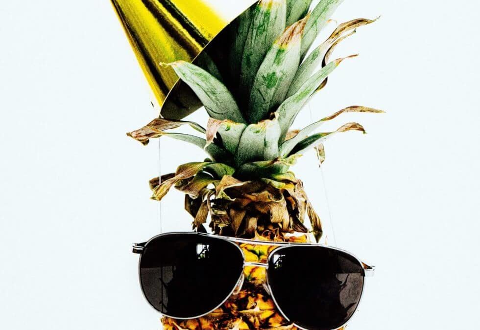 Ananas s naočalama za sunce i zlatnim šeširom za zabave na bijeloj pozadini.